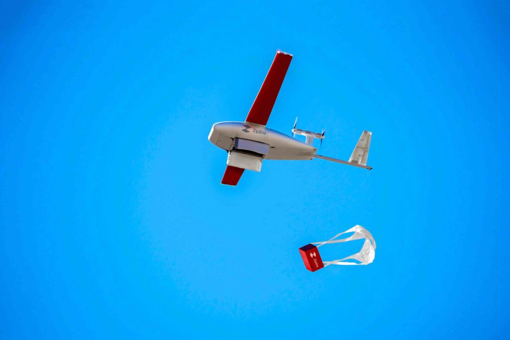 A Zipline drone in the sky with a parachute carrying a package below, demonstrating autonomous delivery services